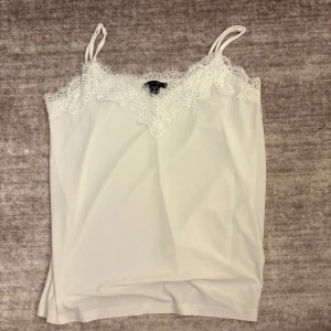 Vit linnetopp med spetsdetaljer - Säljer en vit linnetopp med tunna axelband och vacker spets längs urringningen. Toppen har en mjuk och följsam känsla, perfekt för lager-på-lager eller sommarens varma dagar.