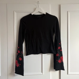 Svart långärmad topp med broderade rosor - Svart långärmad topp med broderade röda rosor på ärmarna från Urban Outfitters. Modellen är croppad och har en rund halsringning.