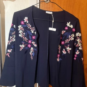 Svart cardigan jacka med broderade blommor - Mörkblå cardigan jacka från Zara med vackra broderade blommor i rosa, vitt och blått på ärmar och framstycke. Jackan är öppen framtill och har lång ärm. Perfekt för att lyfta en enkel outfit.