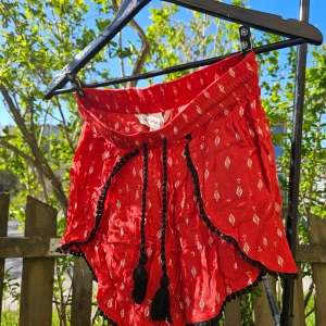 Röda shorts med svarta detaljer - Röda shorts med små vita mönster och svarta tofsar samt dekorativa svarta band längs kanterna. Shortsen har en lös passform och elastisk midja, perfekt för varma dagar.