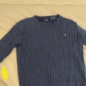 Blå kabelstickad tröja från Gant - Mörkblå kabelstickad tröja från Gant med rund halsringning och diskret logga broderad på bröstet. Klassisk modell med ribbade muddar vid ärmslut och nederkant. Perfekt för en stilren och tidlös look.