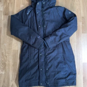 Svart parkajacka från First B - Svart parkajacka från First B med huva och dragkedja framtill. Jackan är lång, har knappar och är fodrad med mjuk fleece på insidan. Perfekt för kyliga dagar och har en klassisk, enkel design.