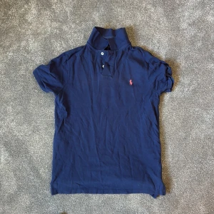 Marinblå pikétröja från Polo Ralph Lauren - Säljer en klassisk marinblå pikétröja från Polo Ralph Lauren med rosa broderad logga på bröstet. Tröjan har korta ärmar, krage och knappar vid halsen. Modellen är custom slim fit och passar perfekt till en avslappnad stil. Liten i Storlek!!!