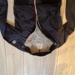 Svart vindjacka från Moncler - Säljer en svart vindjacka från Moncler med rödvit dragkedja och ståkrage. Jackan har långa ärmar, två fickor framtill och Moncler-logga på ena ärmen. Perfekt för dig som vill ha en stilren och sportig look. Jackan är i storlek 158 men sitter som typ en 152.Kan prutas vid snabb affär.