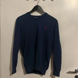 Blå v-ringad tröja från Polo Ralph Lauren - Mörkblå v-ringad tröja från Polo Ralph Lauren med röd broderad logga på bröstet. Långärmad modell i mjukt material med ribbade muddar vid ärmslut och nederkant. Perfekt för lager-på-lager-stil.