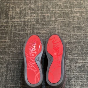 Mörk gråa sneakers med nitar från Christian Louboutin - Säljer ett par mörk gråa sneakers från Christian Louboutin med klassisk röd sula och nitar på tån. Skorna har snörning och är tillverkade i mocka för en lyxig känsla. Perfekta för dig som vill sticka ut med din stil.