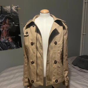 Beige trenchcoat med knappar - Stilren beige trenchcoat med dubbelknäppning och klassisk krage. Jackan har axelklaffar, dekorativa knappar och bälte vid ärmsluten. Perfekt för övergångssäsonger och ger en tidlös look.