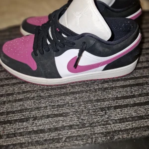 Nike Air Jordan 1 Low rosa och svart - Säljer ett par Nike Air Jordan 1 Low med rosa, svart och vita paneler. Skorna har svart snörning, vit mellansula och klassisk Air Jordan-logga på hälen. Perfekta för dig som vill sticka ut med färg på fötterna.