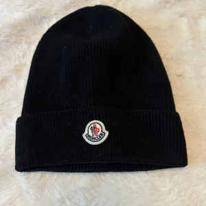 Svart stickad mössa från Moncler - Svart ribbstickad mössa från Moncler med uppvikt kant och klassisk Moncler-logga framtill. Perfekt för kyliga dagar och enkel att matcha med olika outfits. Hör av dig vid frågor