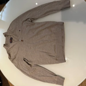 Beige merinoullströja med dragkedja från Morris - Använd fåtal gånger och ser enligt mig helt ny ut. Säljer en stilren beige tröja från Morris i 100% merinoull. Tröjan har dragkedja framtill, ribbade muddar och en liten broderad logga på bröstet. 