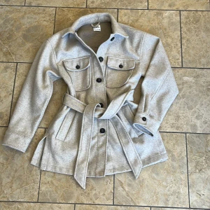 Ljusgrå overshirt med bälte från Zara - Snygg ljusgrå overshirt från Zara med bälte i midjan och stora bröstfickor. Jackan har knappar framtill, långa ärmar och en klassisk krage. Perfekt för lager-på-lager och enkel att styla till olika outfits.