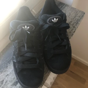 Svarta Adidas Campus sneakers - Säljer ett par svarta Adidas Campus sneakers i storlek 41 med vita detaljer och tre ränder på sidan. Skorna har snörning och är tillverkade i mocka med gummisula. Relativt nya (köpta januari 2025) anledning av att jag säljer de är att jag inte använder de🌸är förhandlingsbar