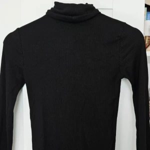 Svart ribbad polotröja - Säljer en svart ribbad polotröja med lång ärm från amisu. Perfekt basplagg med tight passform och hög krage. Enkel att matcha med olika outfits och passar året runt.