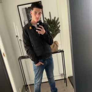 Svart pufferjacka från Parajumper - Säljer en svart vårjacka ish från parajumper med dragkedja och hög krage. Jackan har en gul detalj vid axeln och är lätt vadderad. Perfekt för kyligare dagar och har en stilren look.