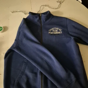Blå zip-jacka från Peak Performance - Mörkblå jacka från Peak Performance med dragkedja och hög krage. Broderad logga på bröstet och två fickor framtill. Perfekt för en sportig stil och enkel att matcha med andra plagg.