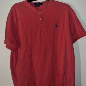 Ralph Lauren - Säljer en rosa t-shirt från Polo Ralph Lauren med tre knappar vid halsen och klassisk broderad logga på bröstet. T-shirten har korta ärmar och är gjord i mjuk bomull. Perfekt för en avslappnad stil.