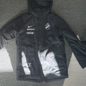 Svart Nike AIK jacka med huva - Svart jacka från Nike med AIK-logga och flera sponsormärken. Jackan har huva, dragkedja och är långärmad. Perfekt för kyligare dagar och sportiga tillfällen.