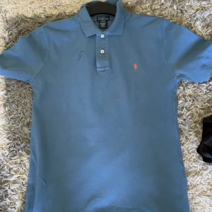 Blå pikétröja från Polo Ralph Lauren - Säljer en klassisk blå pikétröja från Polo Ralph Lauren med orange broderad logga på bröstet. Tröjan har krage och två knappar framtill. Perfekt för en stilren och avslappnad look.