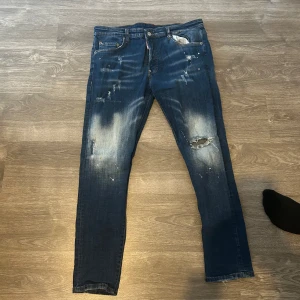 Blå  jeans från Dsquared2 - Säljer ett par blå jeans från Dsquared2 med slitningar, hål och färgstänk. Byxorna har en smal passform och en patch med 'ICON' på baksidan. Klassisk femficksmodell med dragkedja och knapp.
