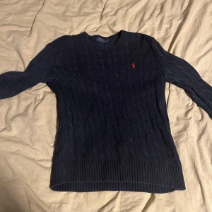 Mörkblå kabelstickad tröja från Polo Ralph Lauren - Säljer en klassisk mörkblå kabelstickad tröja från Polo Ralph Lauren med rund halsringning. Tröjan har lång ärm och den ikoniska röda loggan broderad på bröstet. Perfekt för lager-på-lager och stilrena outfits.