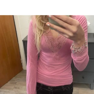 Rosa långärmad topp med spetsdetaljer - Säljer en rosa långärmad topp med v-ringning och fina spetsdetaljer vid halsringningen. Toppen har en tight passform och är perfekt för dig som gillar feminina detaljer.