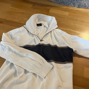 Vit tröja med dragkedja från Massimo Dutti - Vit långärmad tröja från Massimo Dutti med bred mörkblå rand över bröstet och dragkedja vid kragen. Tröjan har ribbade muddar och en hög krage som kan vikas ner. Perfekt för en stilren och avslappnad look.
