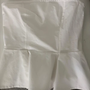 Vit peplum tubtopp - Säljer en stilren vit tubtopp med peplumvolang nertill. Toppen är axelbandslös och har en figurnära passform upptill som släpper ut i en volang. Perfekt för sommardagar eller till festligare tillfällen.