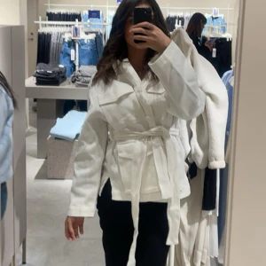 Vit trenchcoat med bälte - Snygg vit trenchcoat med knytskärp i midjan och klassisk krage. Jackan har långa ärmar och en rak passform som ger en stilren look. Perfekt för dig som vill ha en fräsch och trendig jacka till garderoben.