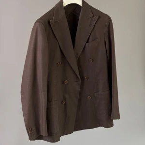 Brun/olivgrön kavaj från Rose & Born - Säljer en brun dubbelknäppt kavaj från Rose & Born, tillverkad i Italien. Kavajen har klassiska slag, två framfickor, bröstficka och dekorativa knappar vid ärmslutet. Perfekt för dig som vill ha en stilren och tidlös look.