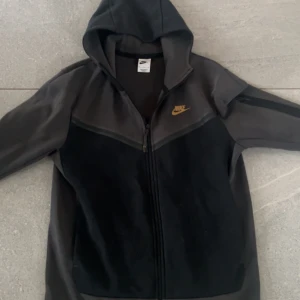 Svart hoodiejacka från Nike - Säljer en svart hoodiejacka från Nike med dragkedja och huva. Jackan har Nike-logga i gult på bröstet och är tillverkad i mjukt material. Perfekt för en sportig och avslappnad stil.