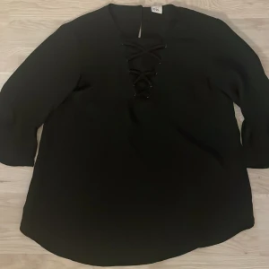 Svart blus med snörning från Lindex - Säljer en svart blus från Lindex med snygg snörning framtill vid halsen. Blusen har långa ärmar och en avslappnad passform, perfekt för en stilren look.