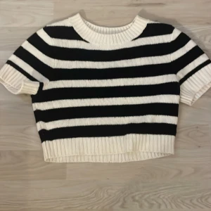 Randig stickad tröja från H&M Divided - Säljer en svartvit, randig stickad tröja från H&M Divided. Tröjan är croppad med korta ärmar och har bred ribbad kant vid hals, ärmslut och nederkant. Perfekt för dig som gillar en trendig och avslappnad stil.