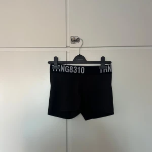 Svarta träningstights shorts från H&M Sport - Svarta korta träningstights från H&M Sport med bred resår i midjan och vit text 'TRNG8310' framtill. Perfekta för gym eller löpning, med tight passform och sportig look.