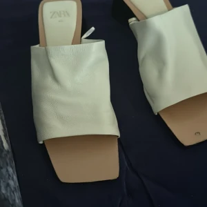 Vita slip-in sandaler från Zara - Säljer ett par trendiga vita slip-in sandaler från Zara med fyrkantig tå och bred rem i mjukt syntetmaterial. Perfekta för sommaren och lätta att matcha till olika outfits.
