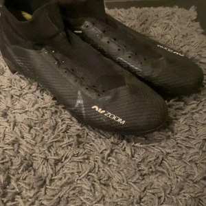 Nike Mercurial Air Zoom fotbollsskor - Säljer ett par svarta Nike Mercurial Air Zoom fotbollsskor med snörning och strumpliknande skaft. Skorna har en lätt ovandel i syntetmaterial och dobbar under sulan för bra grepp på planen. Perfekta för dig som vill ha snabbhet och kontroll.