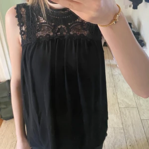 Svart blus med spetsdetaljer - Säljer en svart blus med vackra spetsdetaljer upptill och ärmlös design. Perfekt för dig som vill ha en stilren och elegant topp med fina detaljer vid axlar och halsringning.