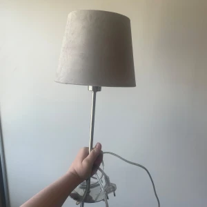 Bordslampa med beige lampskärm - Säljer en stilren bordslampa med beige tygskärm och silverfärgad metallfot. Lampan har en klassisk, rund form och passar perfekt på skrivbord eller nattduksbord. 
