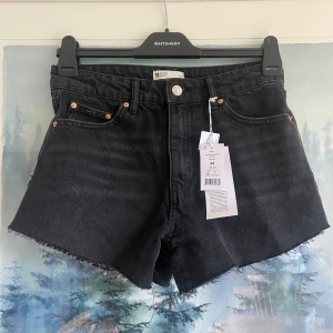 Svarta jeansshorts, stl 34 - Svarta jeansshorts i denim med fransig kant nertill. Klassisk femficksmodell med knapp och dragkedja framtill. Hög midja och raka ben. Perfekta för sommaren och enkel att matcha med olika toppar.