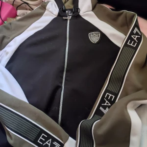 Svart och olivgrön zip-jacka från EA7 Emporio Arman plus tshirt i - Säljer en sportig zip-jacka från EA7 Emporio Armani i svart, olivgrönt och vitt. Jackan har dragkedja framtill, EA7-logga på bröstet och breda loggoremmar längs ärmarna. Perfekt för en streetwear-look. Säljes tillsammans för 250kr