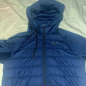 Blå Nike Dri-FIT jacka med huva - Säljer en blå Nike Dri-FIT jacka med huva och dragkedja. Jackan har långa ärmar, quiltade paneler på framsidan och baksidan samt justerbar snörning vid huvan. Perfekt för träning eller vardagsbruk.