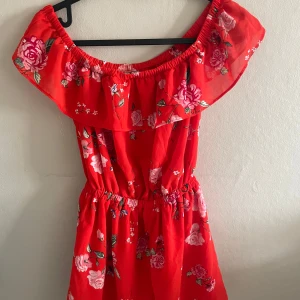 Röd blommig offshoulder kort byxdress från Divided - Säljer en röd offshoulder byxdress från Divided med blommigt mönster och volang upptill. Byxdressen har resår i midjan och är perfekt för varma dagar. Färgglad och somrig stil med fina rosa och vita blommor.