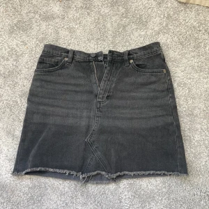 Svart jeanskjol med rå kant - Säljer en svart jeanskjol med rå, fransig nederkant och klassiska femficksdetaljer. Kjolen har dragkedja framtill och en snygg asymmetrisk söm. Perfekt till sneakers eller boots för en cool vardagslook.