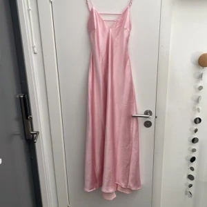 Ljusrosa balklänning i satin  - Supersöt ljusrosa långklänning med smala axelband och v-ringning. Klänningen har en elegant, slät satinliknande yta och en flowy passform. Perfekt för dig som vill ha en feminin och stilren look.