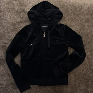 Marinblå  velour hoodie från Juicy Couture - Mysig marinblå hoodie i velour från Juicy Couture med huva, dragkedja och fickor framtill. Klassisk J-detalj på dragkedjan och diskret logga på bröstet. Perfekt för en avslappnad och trendig look.