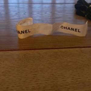 Vit rosett från Chanel - Säljer en vit rosett från Chanel med svart logotyptryck. Perfekt som accessoar till håret eller att knyta runt en väska för en lyxig touch.