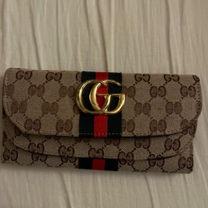 Beige plånbok GG-logga - Säljer en beige plånbok med klassiskt GG-mönster, guldfärgad GG-logga och svart/röd rand på framsidan. Perfekt accessoar för dig som gillar lyxiga detaljer och tidlös design.