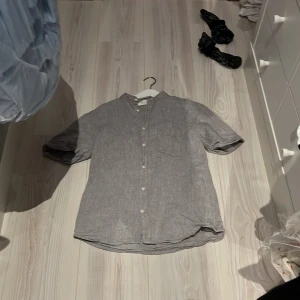 Ljusgrå linneskjorta från H&M - Säljer en ljusgrå skjorta i linne från H&M med ståkrage och vita knappar. Skjortan har korta ärmar och en bröstficka. Perfekt för varma dagar och enkel att matcha med olika outfits.