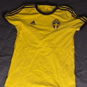 Svenska landslagets fotbollströja från Adidas - Gul kortärmad fotbollströja från Adidas med blå detaljer och Sveriges landslagsemblem på bröstet. Klassisk design med blå ränder på axlarna och Adidas-logga. Perfekt för dig som vill visa ditt stöd för Sverige.