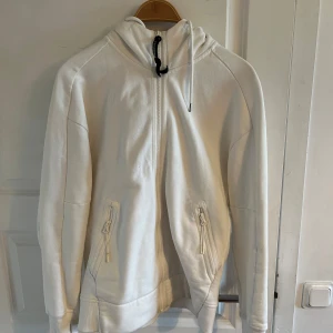 Vit hoodie med goggles från C.P. Company - Säljer en vit hoodie från C.P. Company med dragkedja framtill och två fickor med dragkedja. Huvan har de klassiska goggles-detaljerna. Perfekt för dig som gillar streetwear och vill sticka ut.