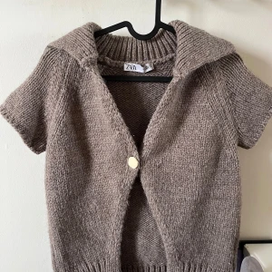 Beige stickad kofta från Zara - Mysig beige stickad kofta från Zara med korta ärmar och stor krage. Koftan har en knapp framtill och ribbad kant vid halsen och nederkanten. Perfekt för lager-på-lager och enkel att styla till olika outfits.